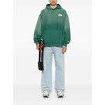 Billionaire Sweaters Green oversized hoodie with front pouch and logo patch, modeled by a person wearing light jeans and sneakers.