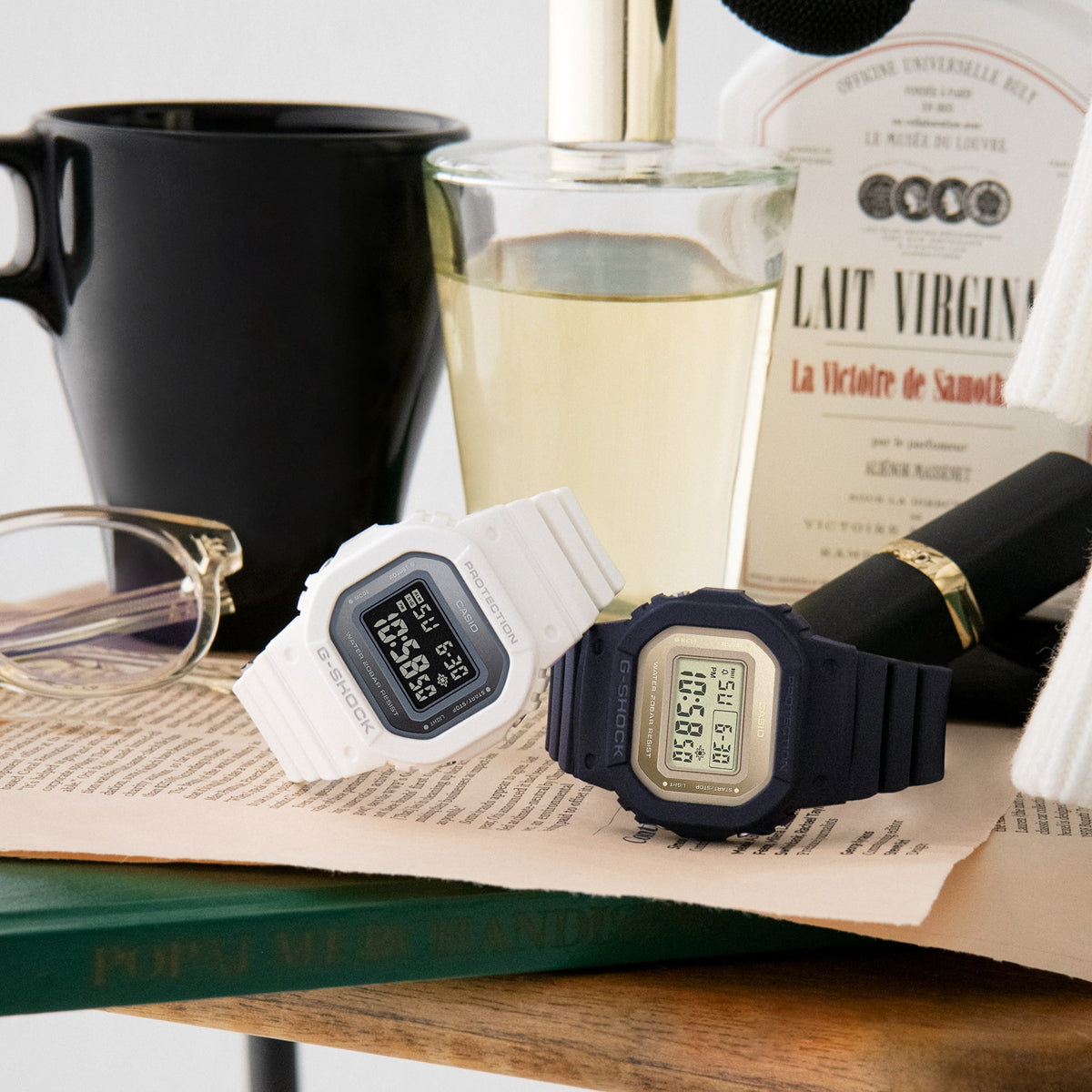 White and black CASIO G-SHOCK Mod. THE ORIGIN Small watches on a table with books and accessories, emphasizing style and versatility.