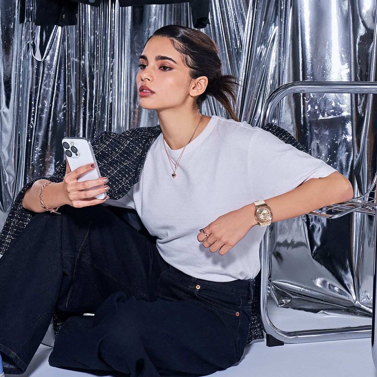 Person wearing CASIO G-SHOCK WATCHES Mod. GM-S2110PG-4AER, holding smartphone, sitting against metallic background.