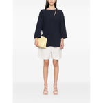 Model wearing Emporio Armani cady blouse in oxford blue, featuring front button fastening and side slits, paired with white shorts and a yellow clutch.