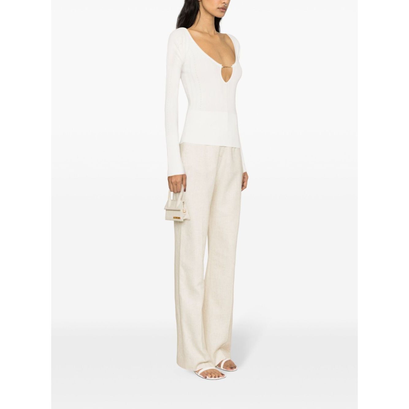Model wearing JACQUEMUS Top White, a long-sleeve off-white ribbed knit with V-neck and gold-tone logo, styled with wide-leg pants.